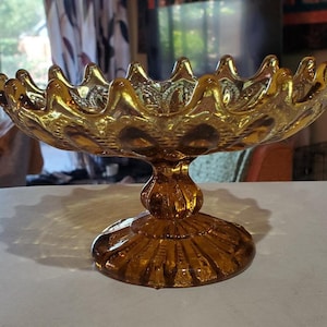 May include: A vintage amber glass pedestal bowl with a scalloped edge and a textured design. The bowl is on a pedestal base with a fluted design.