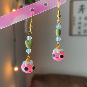 May include: A pair of pink and white clay earrings with a green heart and blue beads hanging from each earring.