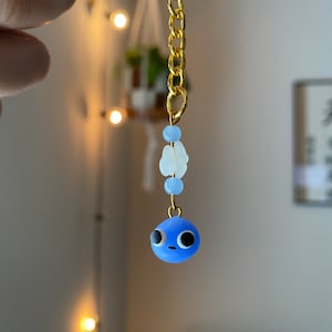 May include: A handmade keychain featuring a blue character with large eyes. The keychain has a gold-colored chain and ring, with light blue and white beads. The character is a small, round, blue figure with black eyes.