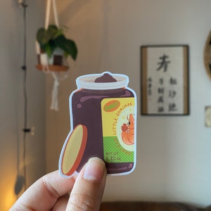 May include: A sticker of a jar of brown sauce with a yellow label that says "Little Shrimps" and "Sauteed" with a cartoon shrimp on the label. The label also says "Net Wt. 8.8 oz".