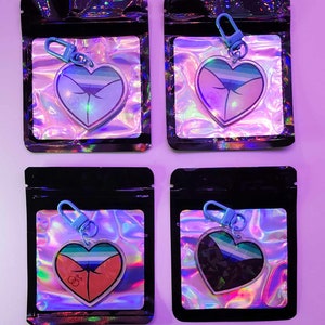 May include: Four clear heart-shaped keychains with different colored designs. Each keychain is packaged in a black resealable bag with a holographic iridescent background. The keychains feature a blue, red, and black design with a rainbow stripe.