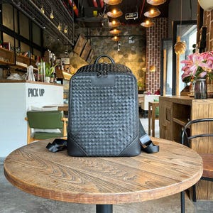 May include: A black backpack with a woven design sits on a round wooden table. The backpack has a top handle, a front pocket, and adjustable shoulder straps. The background shows a cafe setting with a "Pick Up" sign.