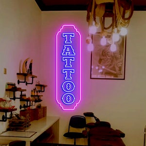 Puede incluir: Un letrero de ne&oacute;n con la palabra "TATTOO" en letras azules sobre un fondo rosa. El letrero est&aacute; colgado en una pared blanca en una habitaci&oacute;n con una silla de cuero marr&oacute;n y una silla negra.