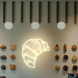 May include: A wall display featuring a glowing neon croissant sign and various miniature baked goods. The display includes round, white globe lights and wooden accents, creating a warm and inviting atmosphere. The baked goods are in various shapes and colors.