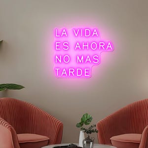 Könnte beinhalten: Ein leuchtend rosa Neon-Schild mit dem spanischen Text "LA VIDA ES AHORA NO MAS TARDE" an einer neutralen Wand. Das Schild hat eine besondere Form. Der Raum zeigt zwei korallenfarbene Sessel.