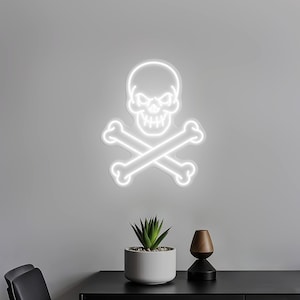 Può includere: Un'insegna al neon bianca a forma di teschio e ossa incrociate è montata su un muro grigio. Sotto l'insegna, una piccola pianta succulenta in un vaso bianco e un vaso decorativo in legno.
