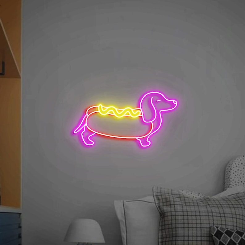 Op de afbeelding: Een neonbord in de vorm van een teckelhond die een hotdogkostuum draagt. De hond is roze en de hotdog is geel met mosterd.