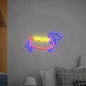 Op de afbeelding: Neon-bord in de vorm van een teckel die een hotdog-kostuum draagt. Het bord is blauw, rood en geel.
