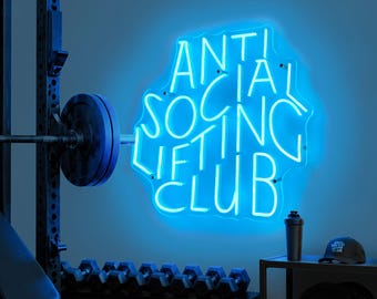 May include: A blue neon sign that reads "ANTI SOCIAL LIFTING CLUB" hangs in a gym setting. The sign is surrounded by gym equipment, including a weight bench, barbell, dumbbells, and weight plates. A black shaker bottle and a cap are on a shelf.