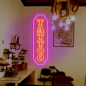 Puede incluir: Un letrero de ne&oacute;n con la palabra "TATTOO" en letras rojas con un contorno blanco. El letrero est&aacute; iluminado con un brillo rosa y est&aacute; colgado en una pared blanca.