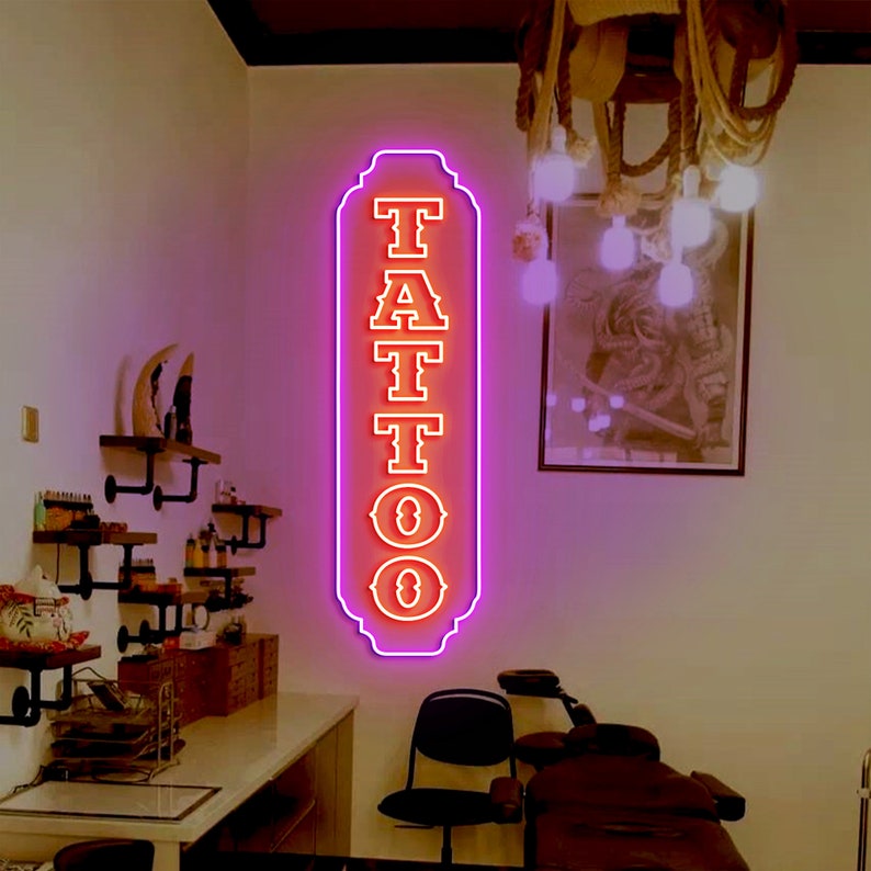 Puede incluir: Un letrero de ne&oacute;n con la palabra "TATTOO" en letras rojas con un contorno blanco. El letrero est&aacute; iluminado con un brillo rosa y est&aacute; colgado en una pared blanca.