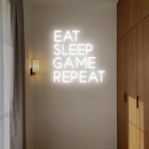 Könnte beinhalten: Weißes Neon-Schild an einer Wand mit der Aufschrift "EAT SLEEP GAME REPEAT". Das Schild ist beleuchtet und an einer hellen Wand angebracht. Links vom Schild befindet sich eine kleine Wandleuchte, rechts ein Holzschrank.