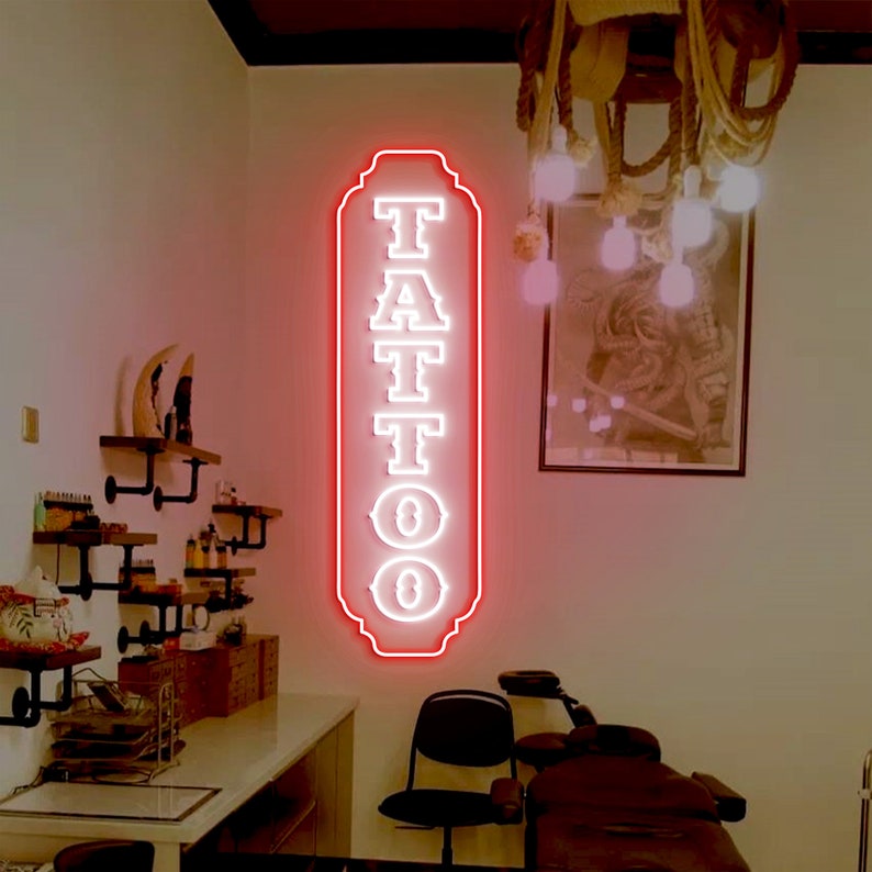 Puede incluir: Un letrero de ne&oacute;n rojo con letras blancas que dice "TATTOO" en una fuente de estilo vintage. El letrero est&aacute; colgado en una pared blanca en una habitaci&oacute;n con una silla de tatuaje y otros muebles.