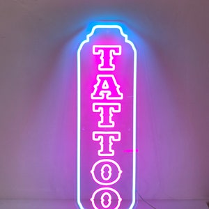 Puede incluir: Un letrero de ne&oacute;n con un letrero "TATTOO" rosa con contornos blancos. El letrero tiene forma rectangular con esquinas redondeadas y un borde decorativo. El letrero est&aacute; iluminado con un brillo azul alrededor de los bordes.