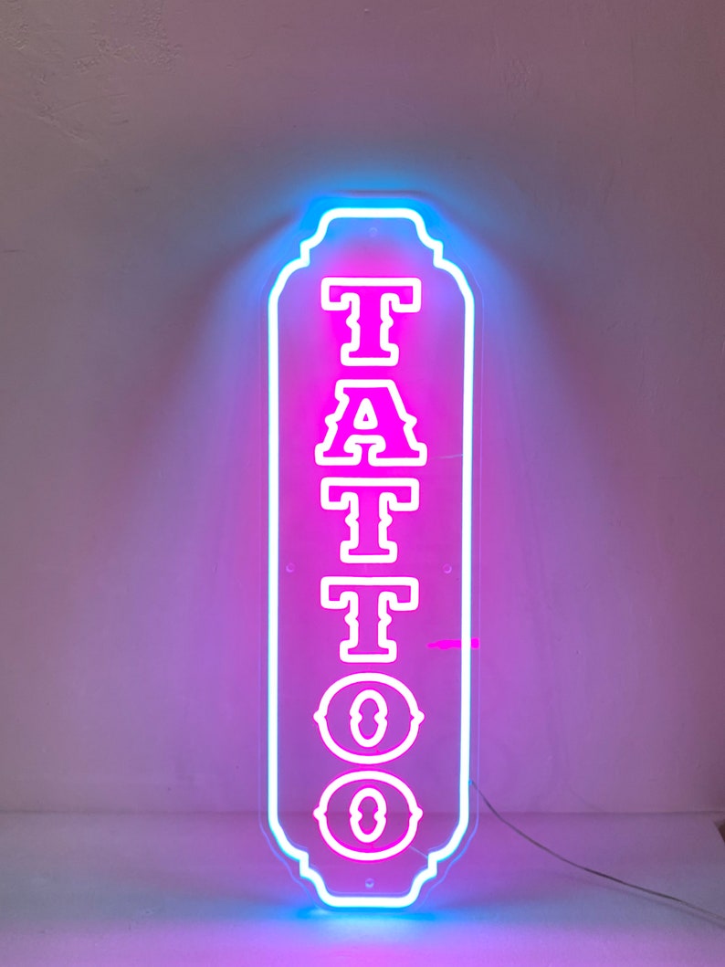 Puede incluir: Un letrero de ne&oacute;n con un letrero "TATTOO" rosa con contornos blancos. El letrero tiene forma rectangular con esquinas redondeadas y un borde decorativo. El letrero est&aacute; iluminado con un brillo azul alrededor de los bordes.