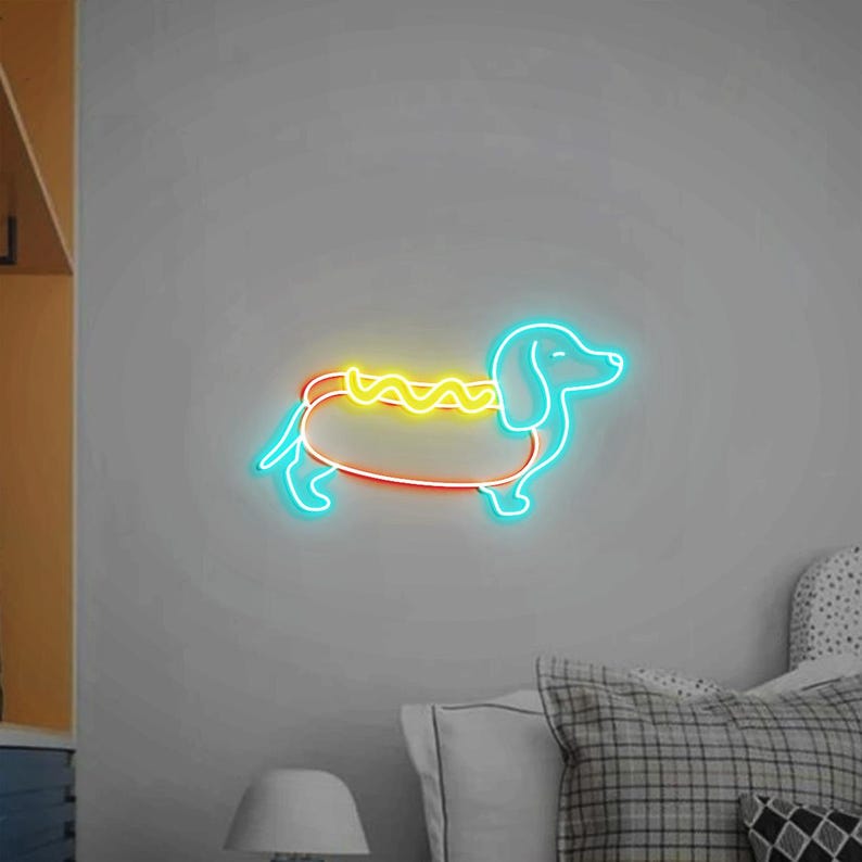 Op de afbeelding: Een neon-bord in de vorm van een teckel die een hotdog-kostuum draagt. De hotdog is geel met een mosterdlijn en de teckel is blauw.