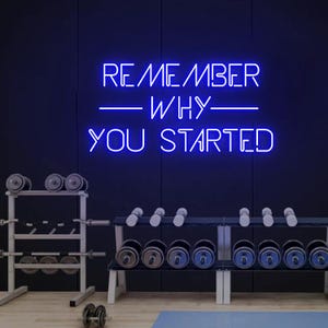 Könnte beinhalten: Blaues Neon-Schild mit dem Text "Remember Why You Started" an einer schwarzen Wand in einem Fitnessstudio mit Hantelständern und Kurzhanteln.