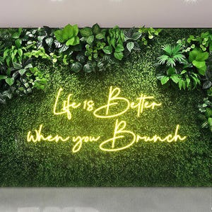 May include: A wall covered in green foliage with a neon sign. The sign reads "Life is Better when you Brunch" in a cursive font. The neon sign glows with a warm light, creating a cheerful atmosphere. The wall is a backdrop for events.