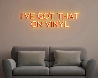 May include: Orange neon sign with the text "I've got that on vinyl" on a beige wall above a gray couch with two pillows.