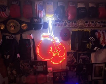 May include: Neon sign of two red boxing gloves hanging from a white string. The sign is mounted on a wall with boxing memorabilia, including boxing gloves, belts, and photos.