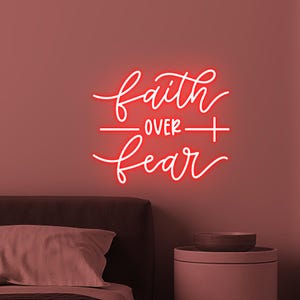 Könnte beinhalten: Ein rotes Neon-Schild mit den Worten "faith over fear" in einer Schreibschrift. Das Schild ist an einer rosa Wand über einem Bett mit einem Kissen und einem Beistelltisch mit einer Schüssel angebracht.