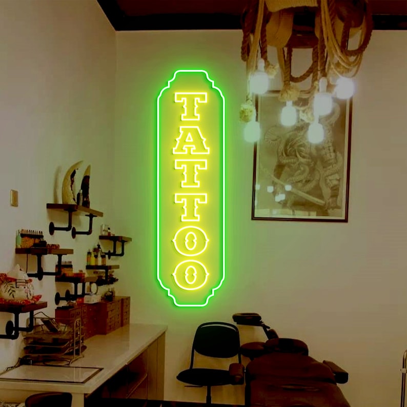Puede incluir: Un letrero de ne&oacute;n con contorno verde y letras amarillas que dice "TATTOO". El letrero est&aacute; montado en una pared blanca en una habitaci&oacute;n con una mesa de madera y una silla negra.