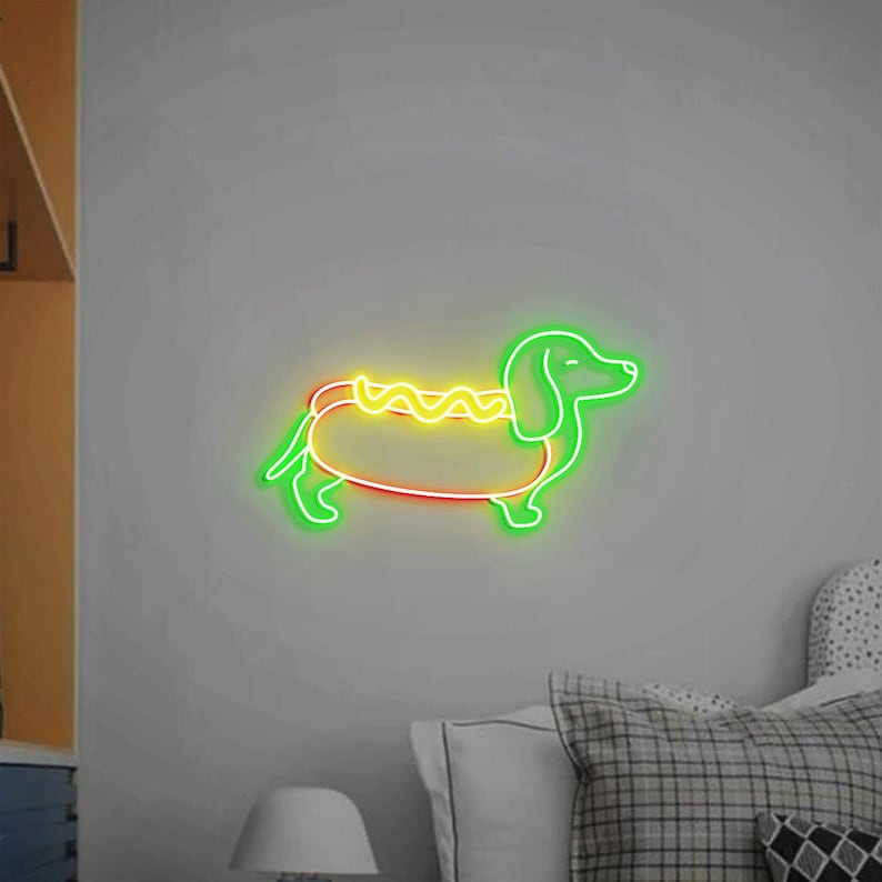 Op de afbeelding: Een neonbord in de vorm van een teckel die een hotdogkostuum draagt. De hond is groen, de hotdog is geel en de mosterd is geel.