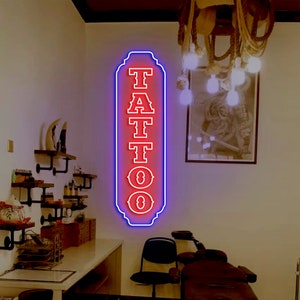 Puede incluir: Un letrero de ne&oacute;n rojo con la palabra "TATTOO" en una fuente de estilo vintage. El letrero est&aacute; bordeado de azul y tiene un borde decorativo. El letrero est&aacute; colgado en una pared blanca en un sal&oacute;n de tatuajes.