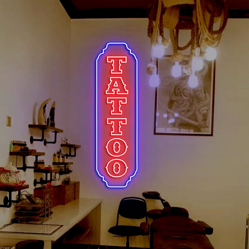 Puede incluir: Un letrero de ne&oacute;n rojo con la palabra "TATTOO" en una fuente de estilo vintage. El letrero est&aacute; bordeado de azul y tiene un borde decorativo. El letrero est&aacute; colgado en una pared blanca en un sal&oacute;n de tatuajes.