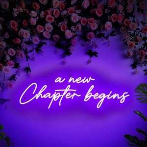 May include: A neon sign with the words "a new Chapter begins" in white script against a purple background. The sign is surrounded by pink and white roses and greenery, creating a romantic and celebratory atmosphere.