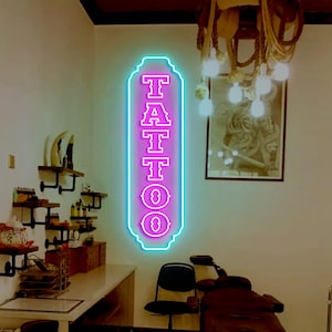Puede incluir: Un letrero de neón con la palabra "TATTOO" en letras rosas con un contorno azul. El letrero está colgado en una pared blanca en una habitación con una silla de tatuaje y otros muebles.