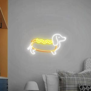 Op de afbeelding: Een wit neonbord in de vorm van een teckel met een gele neonhotdog op zijn rug. Het bord is gemonteerd op een witte muur.