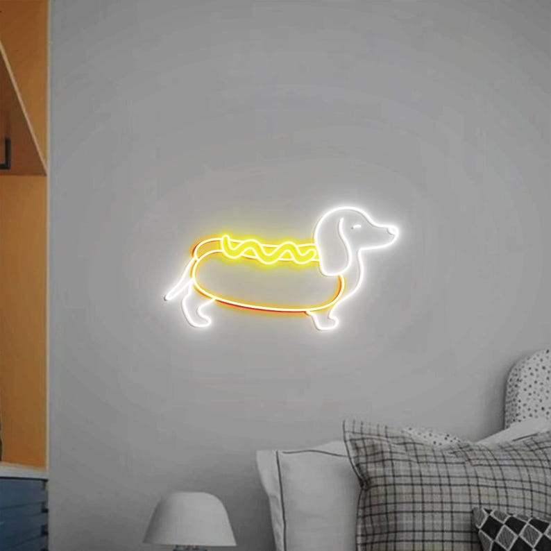 Op de afbeelding: Een wit neonbord in de vorm van een teckel met een gele neonhotdog op zijn rug. Het bord is gemonteerd op een witte muur.