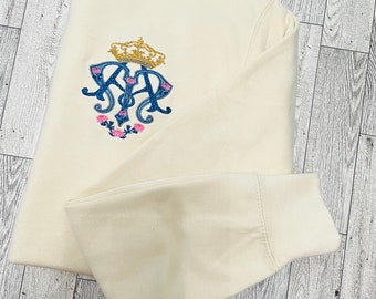May include: A cream colored sweatshirt with a blue and gold embroidered logo featuring a crown, the letters "K" and "A", and pink roses.