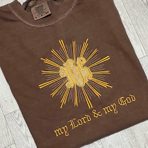 May include: A brown t-shirt with a gold embroidered design of a sunburst with a monogram in the center. The text "my Lord & my God" is embroidered below the design.