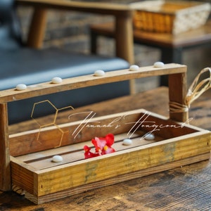 May include: A wooden tray with a handle, featuring a honeycomb design and white ceramic accents. A single red flower is placed inside the tray.