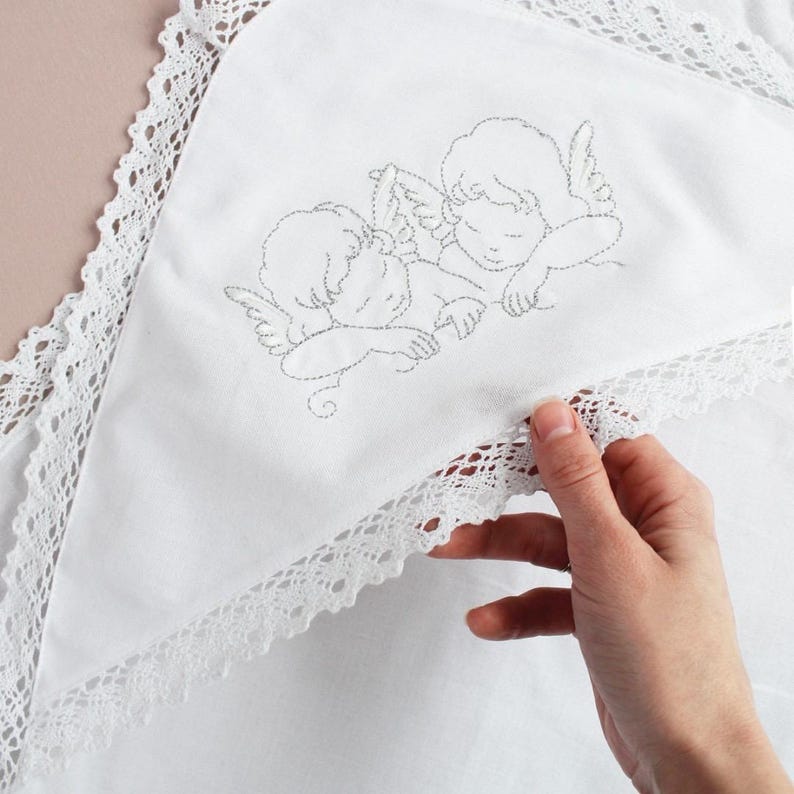 May include: White baby blanket with a delicate lace trim and embroidered angel design. The blanket features a soft, textured surface and a detailed depiction of two cherubs. The lace trim adds a touch of elegance to this baby item.
