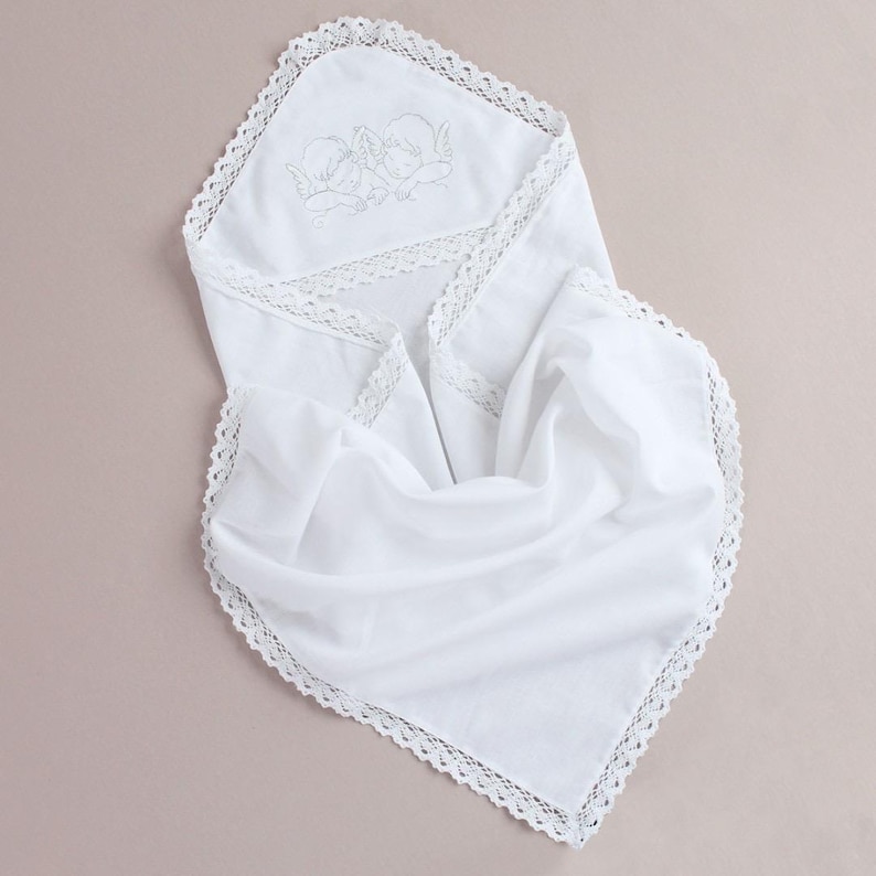 May include: White hooded baby blanket with embroidered angel design and delicate lace trim. The blanket is made of soft fabric and is folded, showcasing the hood and lace details. The overall aesthetic is delicate and suitable for a baby.