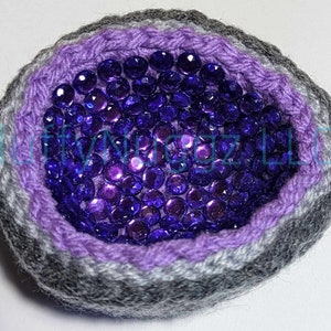 Op de afbeelding: Een gehaakte amethistgeode met een gestreepte buitenkant in paars en grijs en een centrum gevuld met paarse, iriserende pailletten.