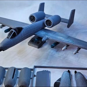 May include: A detailed model of a gray A-10 Thunderbolt II aircraft, featuring twin engines, wings with mounted missiles, and a detailed cockpit. Additional unassembled parts are visible below the plane. The model is likely for display or hobby purposes.