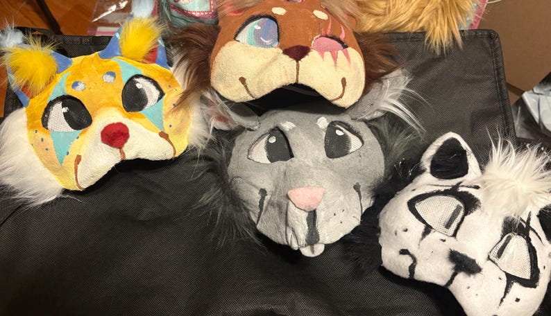 Op de afbeelding: Vier dierenmaskers met gestileerde ontwerpen. E&eacute;n masker is geel met blauwe accenten en geel bont, &eacute;&eacute;n is bruin met donker bont, &eacute;&eacute;n is grijs met donker bont en &eacute;&eacute;n is wit met zwarte markeringen. Elk masker heeft gaas over de ogen.
