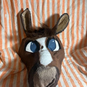 Op de afbeelding: Een bruin en wit dierenkopmasker met grote blauwe ogen met zwarte omlijning, hoge puntige oren en een pluk donkerbruin haar op het voorhoofd. Het masker heeft een witte bles die in het midden over het gezicht naar de snuit toe loopt.