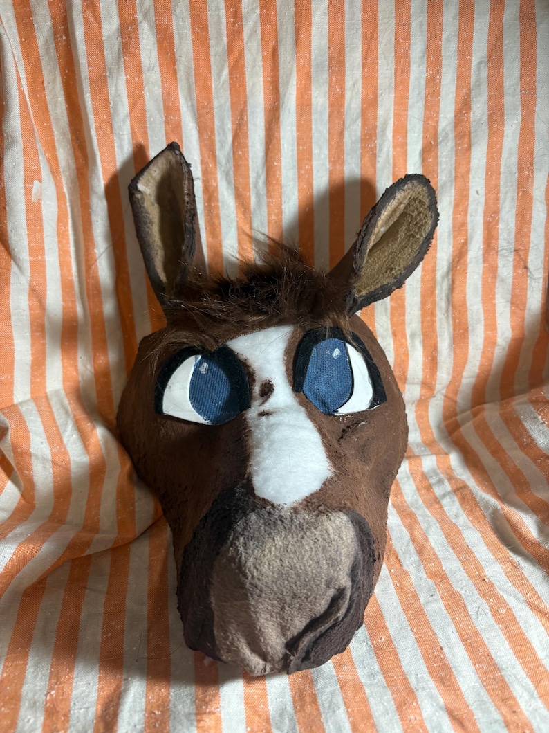 Op de afbeelding: Een bruin en wit dierenkopmasker met grote blauwe ogen met zwarte omlijning, hoge puntige oren en een pluk donkerbruin haar op het voorhoofd. Het masker heeft een witte bles die in het midden over het gezicht naar de snuit toe loopt.