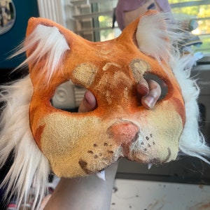 Op de afbeelding: Een kattenmasker gemaakt van oranje en beige viltmateriaal, voorzien van witte imitatiebontplukjes op de oren en zijkanten. Het masker heeft twee oogopeningen, een roze neus en donkerbruine snorharen. Het oppervlak heeft een textuur.