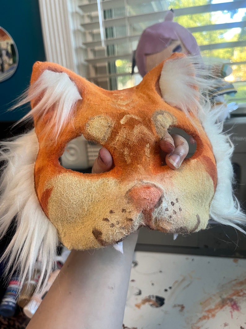 Op de afbeelding: Een kattenmasker gemaakt van oranje en beige viltmateriaal, voorzien van witte imitatiebontplukjes op de oren en zijkanten. Het masker heeft twee oogopeningen, een roze neus en donkerbruine snorharen. Het oppervlak heeft een textuur.