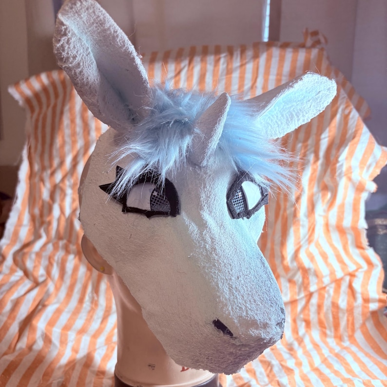 Op de afbeelding: Een wit eenhoornmasker met een getextureerd oppervlak, een centrale hoorn en lichtblauwe plukjes imitatiebont tussen de oren. Het masker heeft zwart omrande oogopeningen met gaas en een puntige snuit, getoond op een neutraal paspophoofd.