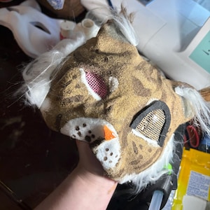 Op de afbeelding: Een handgemaakt katachtig masker met een bruin en beige getextureerd oppervlak met donkerbruine vlekken. Het masker bevat witte imitatiebontaccenten, een kleine oranje neus en twee oogopeningen bedekt met roze en gouden gaasschermen.