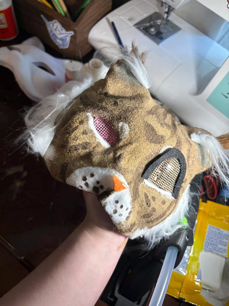 Op de afbeelding: Een handgemaakt katachtig masker met een bruin en beige getextureerd oppervlak met donkerbruine vlekken. Het masker bevat witte imitatiebontaccenten, een kleine oranje neus en twee oogopeningen bedekt met roze en gouden gaasschermen.