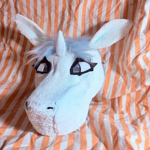 Op de afbeelding: Een wit eenhoornmasker met een textuur, een centrale hoorn en lichtblauw imitatiebont. Het masker heeft grote ogen met zwarte omlijningen en gaasvensters voor het zicht, evenals twee rechtopstaande oren met lichtblauwe accenten aan de binnenkant.