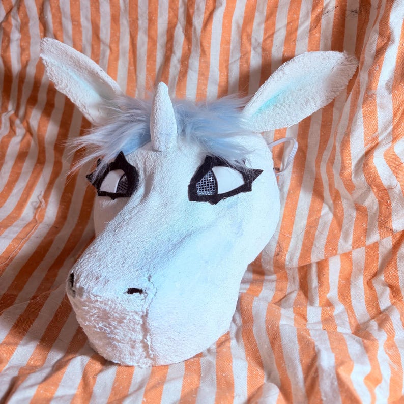 Op de afbeelding: Een wit eenhoornmasker met een textuur, een centrale hoorn en lichtblauw imitatiebont. Het masker heeft grote ogen met zwarte omlijningen en gaasvensters voor het zicht, evenals twee rechtopstaande oren met lichtblauwe accenten aan de binnenkant.
